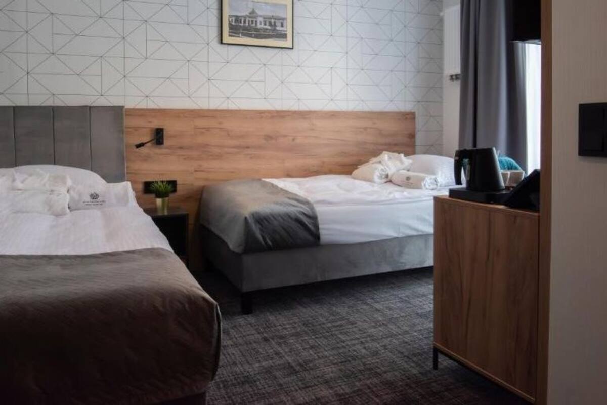 A hotel room with two beds, a wooden headboard, a small bedside table with a lamp, and a view of a building through the window.