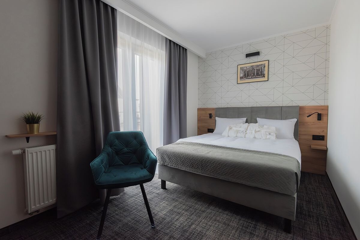 A hotel room with a bed, a chair, a radiator, a window with curtains, and a picture on the wall.