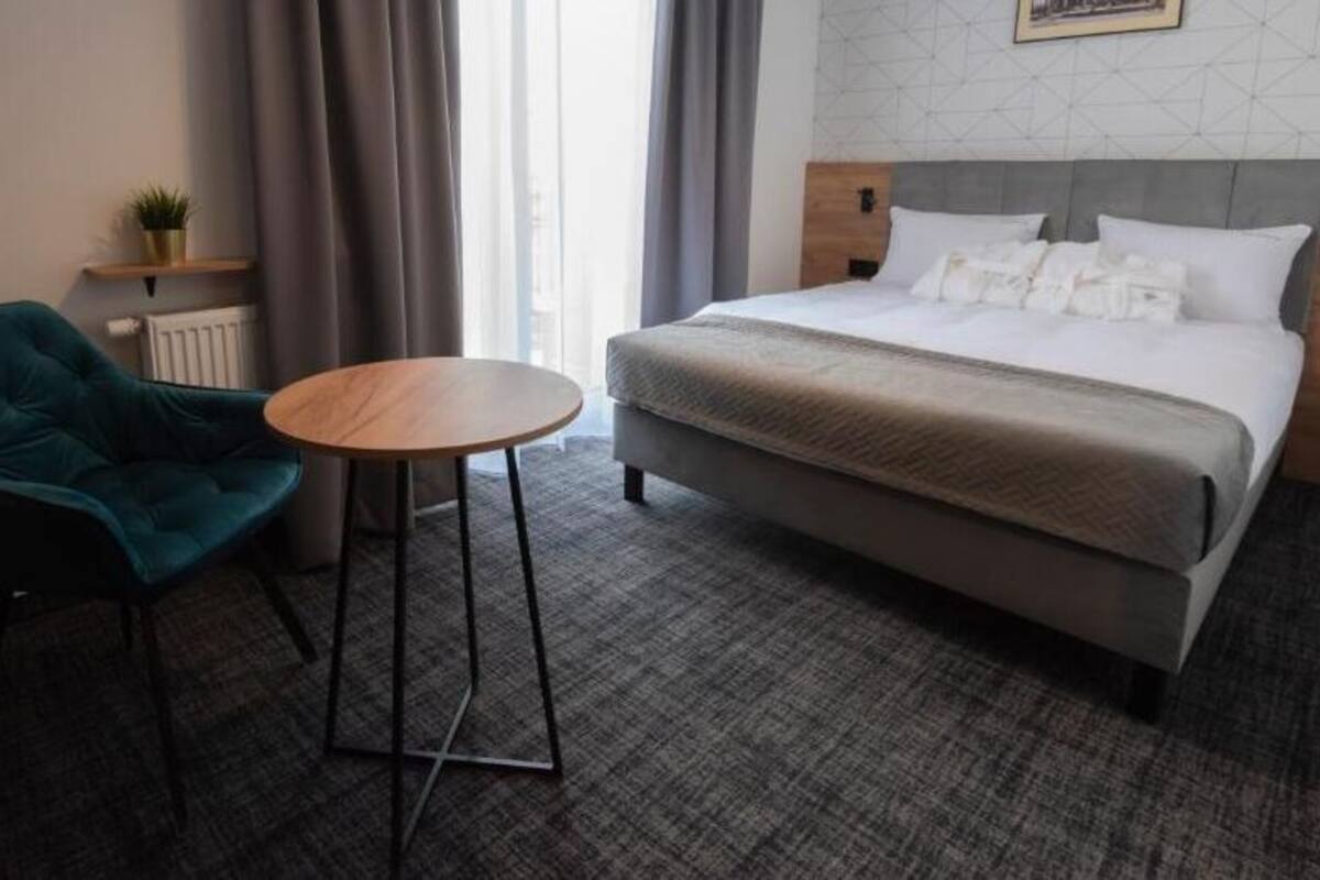 A hotel room with a bed, a teal armchair, a small round table, and a radiator.