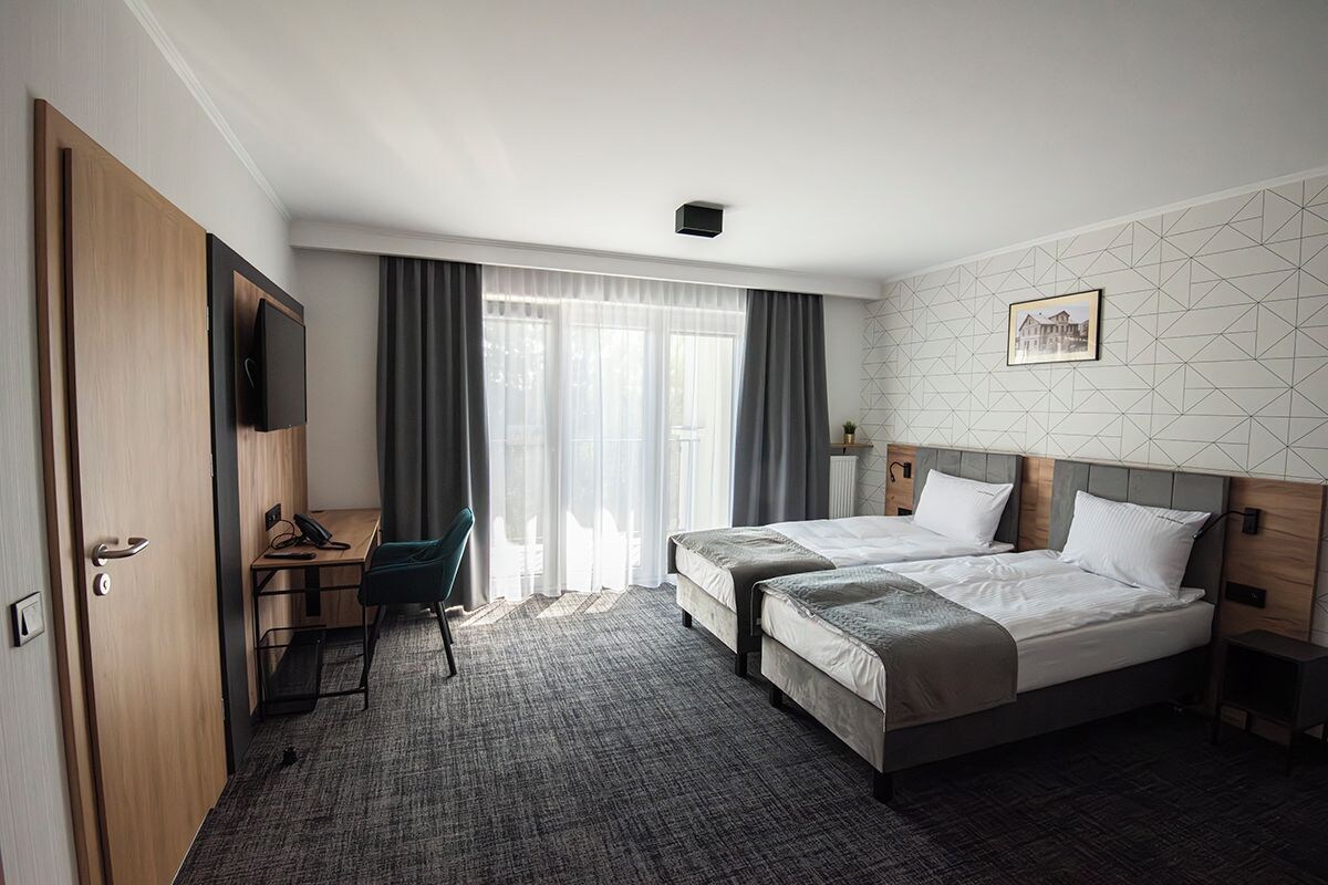 A hotel room with two beds, a desk with a chair, a television, and a large window with curtains.
