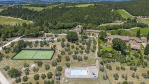 Eine Vogelperspektive auf eine ländliche Gegend mit einem Tennisplatz, einem Schwimmbad und Steinbauten inmitten bewaldeter Hügel.