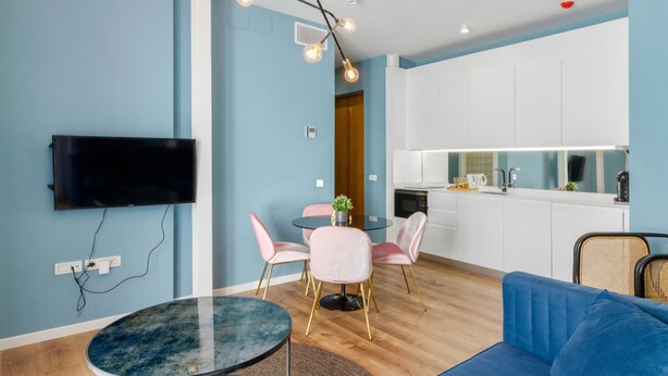 Ein modernes Wohnzimmer mit einem blauen Sofa, einem Glastisch und einem an der Wand montierten Fernseher. Es gibt eine Küchenzeile mit weißen Schränken und einer Mikrowelle.