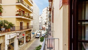 Une rue étroite bordée d’immeubles à plusieurs étages, de balcons ornés de plantes en pot et de devantures de magasins.