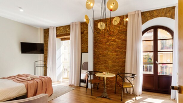 Ein modernes Hotelzimmer mit Backsteinwand, Holzboden, Bett, Fernseher, Esstisch mit Stühlen und einem großen Fenster mit Vorhängen.