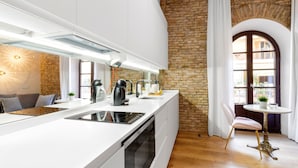 Una cocina moderna con encimera blanca, horno integrado y cafetera. Se observa una pared de ladrillo y una ventana con cortinas.