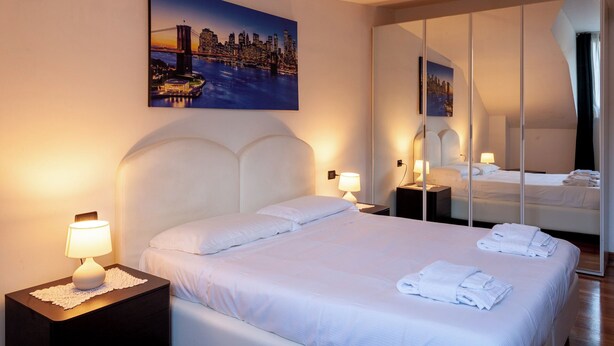 A hotel room with a large bed, two bedside tables with lamps, a cityscape painting, and a full-length mirror. - BnButler - Asiago 41 (Milan)