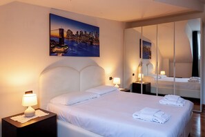 A hotel room with a large bed, two bedside tables with lamps, a cityscape painting, and a full-length mirror. - BnButler - Asiago 41 (Milan)