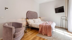 Ein modernes Schlafzimmer mit einem gebogenen Sessel, einem Bett mit rosa Decke, einem Nachttisch und einem wandmontierten Fernseher.