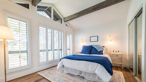 Une chambre avec un grand lit, des murs blancs et un plancher en bois. On y trouve des fenêtres avec des stores, une table de chevet avec une lampe et un tableau accroché au mur.