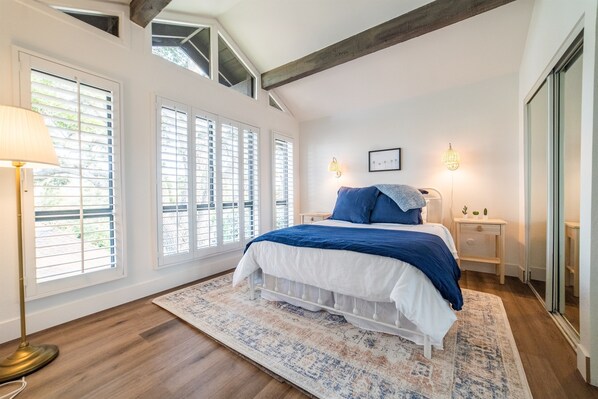 Ein Schlafzimmer mit einem großen Bett, weißen Wänden und Holzboden. Es gibt Fenster mit Jalousien, einen Nachttisch mit Lampe und ein Bild an der Wand.