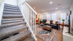 Un escalier avec des marches recouvertes de moquette et des rampes blanches, menant à un salon comprenant un canapé, une table basse et un tapis à motifs.