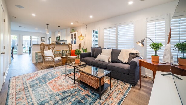 Un salon moderne avec un canapé gris foncé, une table basse en verre et un tapis à motifs. On y trouve également un fauteuil en osier et une petite table avec une lampe. La pièce est dotée de grandes fenêtres équipées de stores et offre une vue sur le jardin.