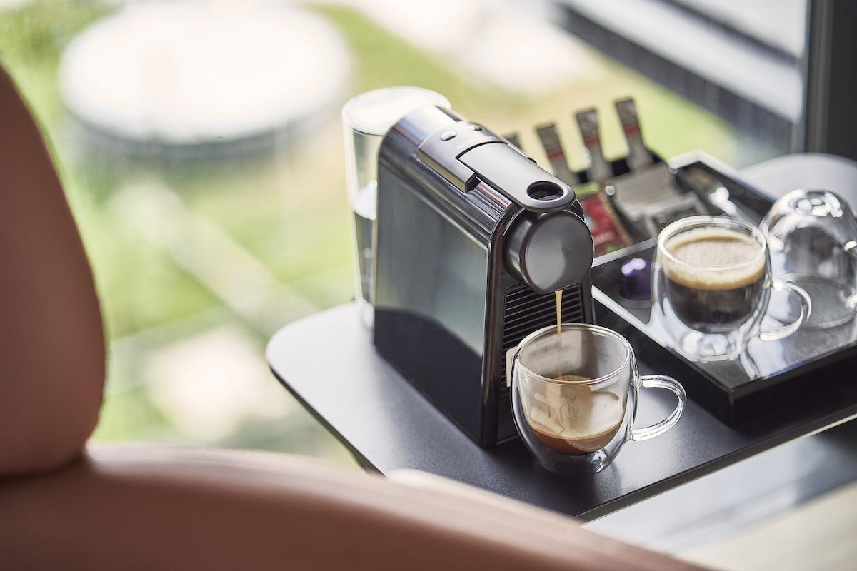 Eine Kaffeemaschine auf einem Tablett mit zwei Kaffeetassen, einem Glas Wasser und einer kleinen Flasche, vermutlich für Zusätze oder Sirups.