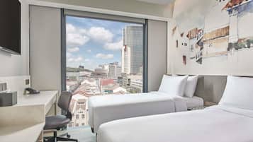 Habitación de hotel con escritorio, dos camas, una silla y un ventanal con vistas a la ciudad.