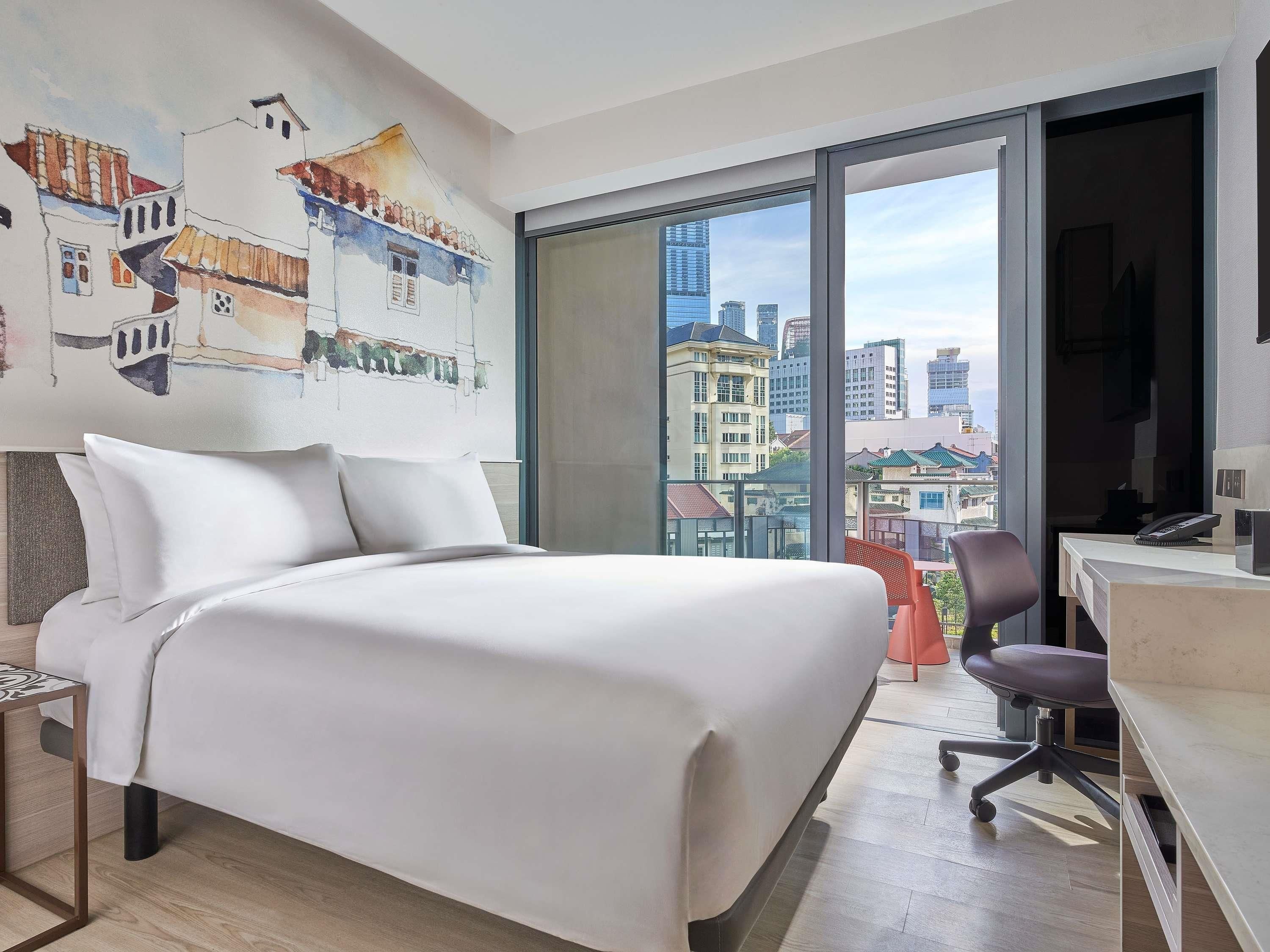 A modern hotel room with a large bed, a desk, and a view of the cityscape through a large window.