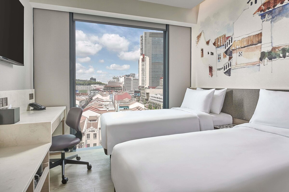 A hotel room with a desk, two beds, a chair, and a large window with a city view.