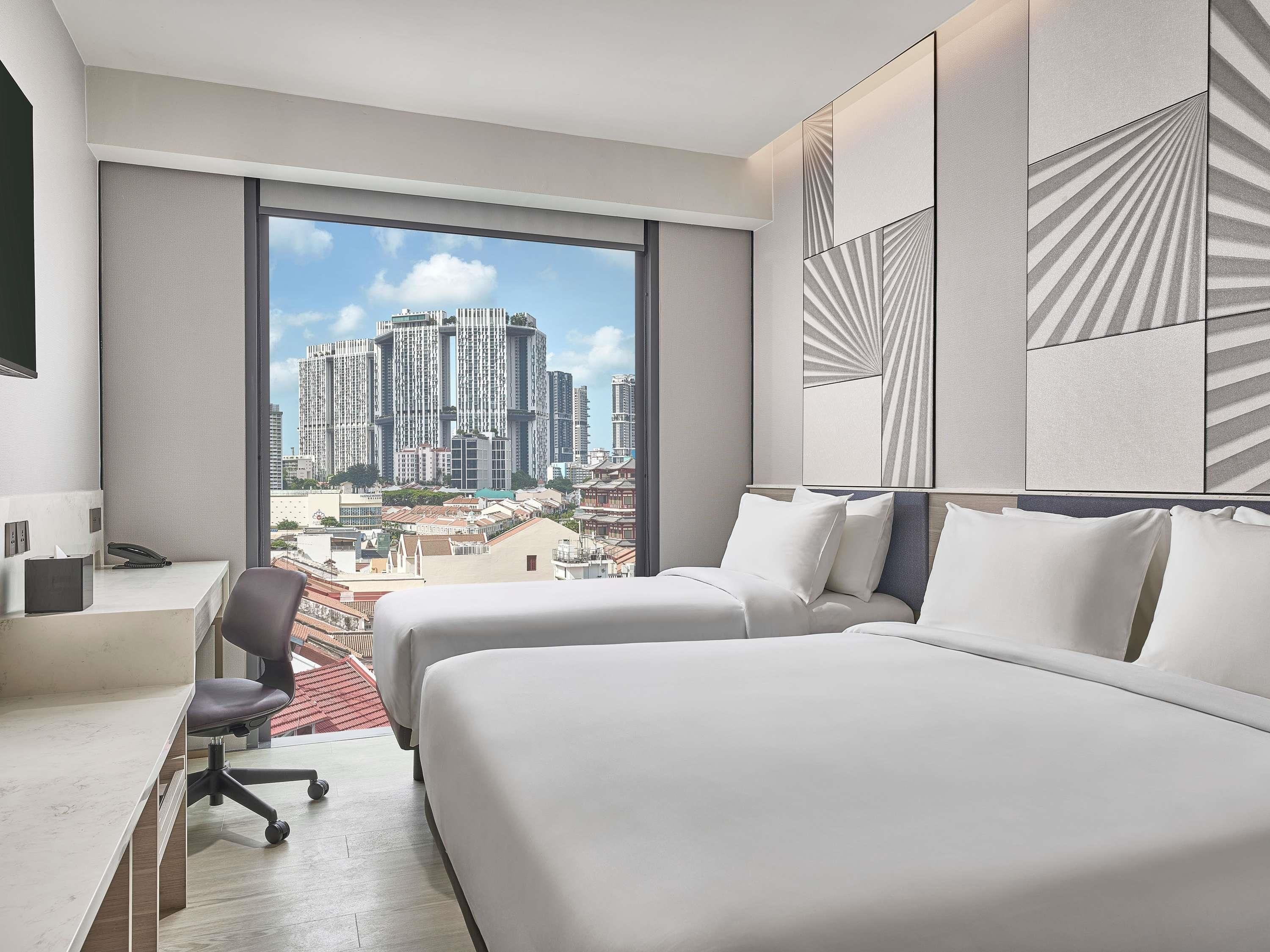 A hotel room with two beds, a desk, and a chair. There is a large window with a city view.