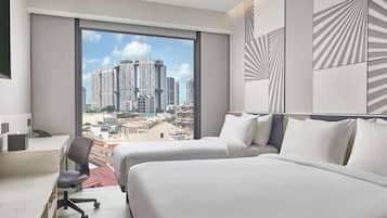 Ein Hotelzimmer mit zwei Betten, einem Schreibtisch und einem Sessel. Es gibt ein großes Fenster mit Blick auf die Stadt.