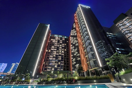 A modern hotel with a pool, surrounded by high-rise buildings and illuminated at night.