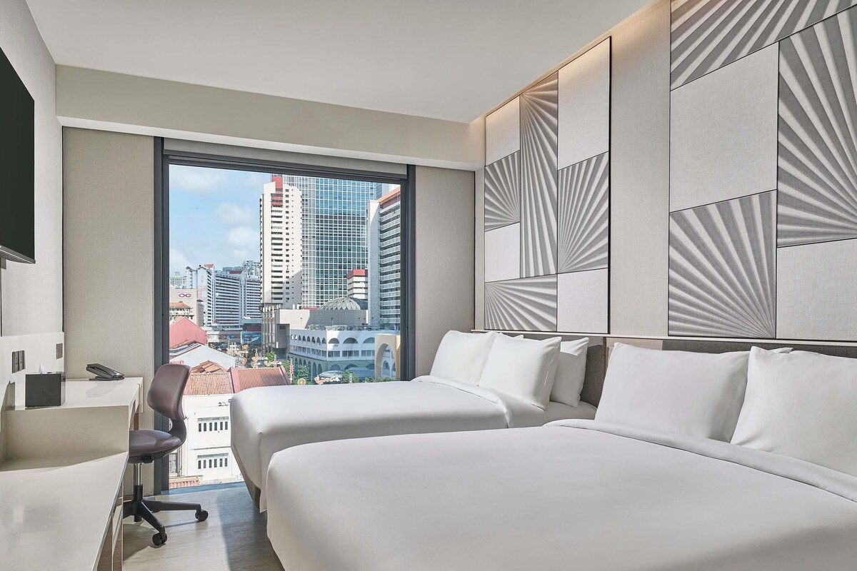 A hotel room with two beds, a desk with a chair, and a large window offering a city view.