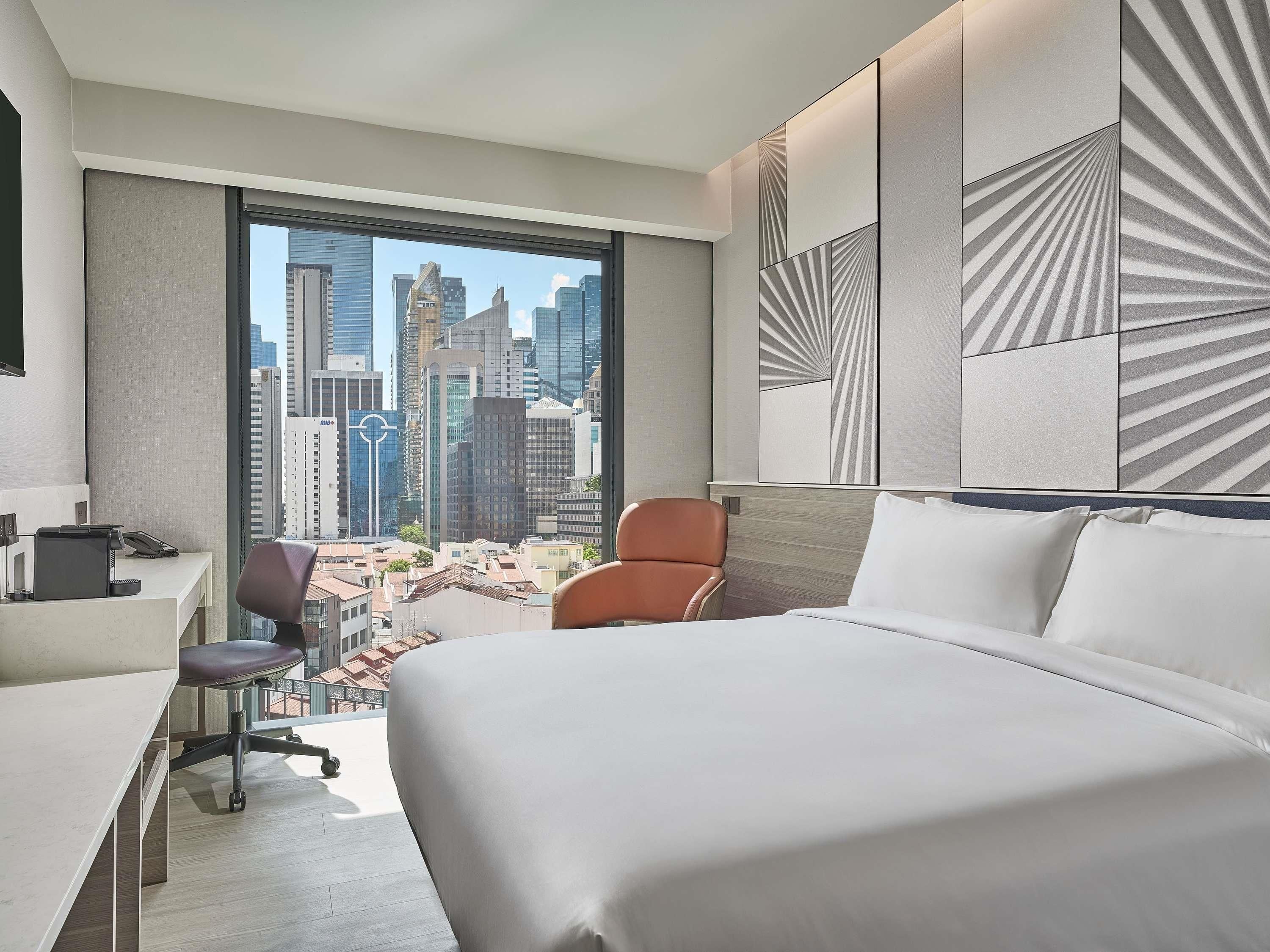 Une chambre d’hôtel avec un grand lit, un bureau et une chaise. Elle offre une vue sur la ville grâce à une grande fenêtre.