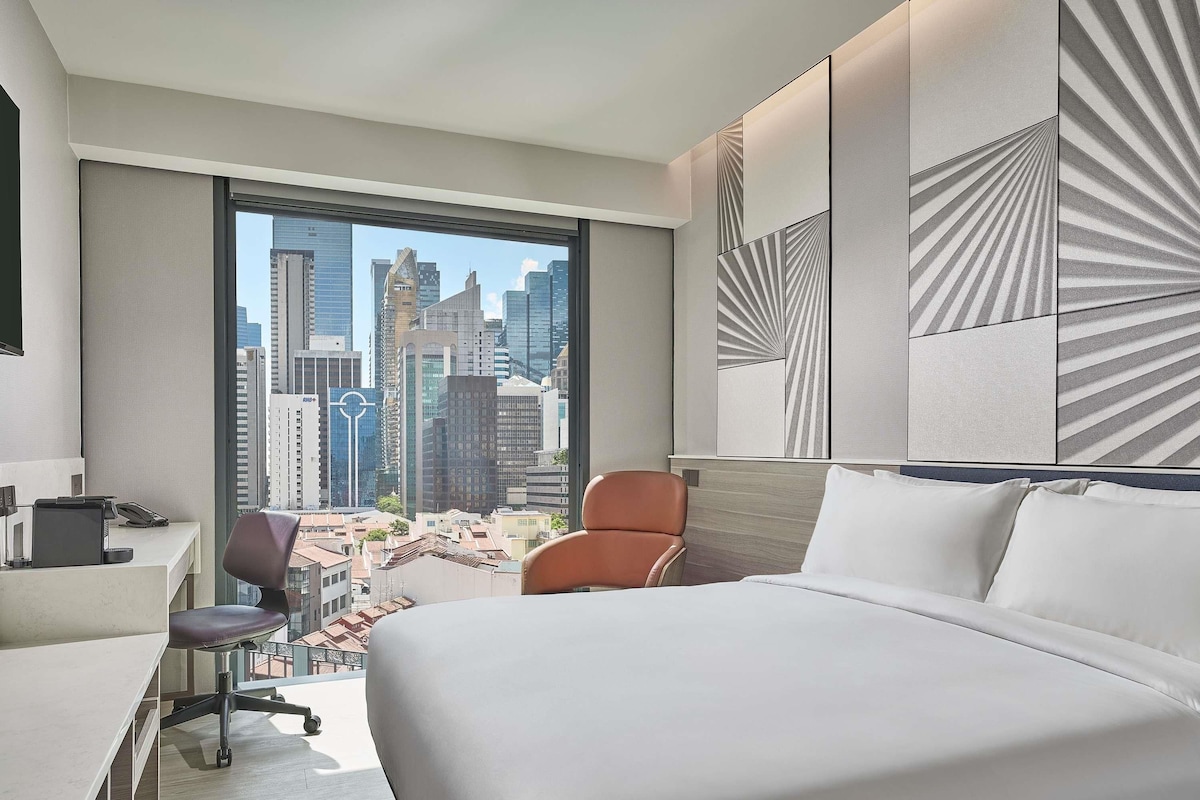 A hotel room with a large bed, a desk, and a chair. It features a city view through a large window.