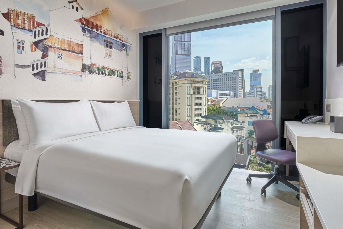 A hotel room with a large bed, a desk, and a view of a cityscape.
