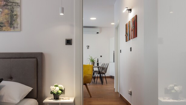 Een moderne slaapkamer met een bed, nachtkastje en een kleine plant. Een gang leidt naar een eetgedeelte met een tafel en stoelen.