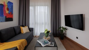 Een moderne woonkamer met een grijze bank, een salontafel met tijdschriften, een televisie en een schilderij aan de muur.