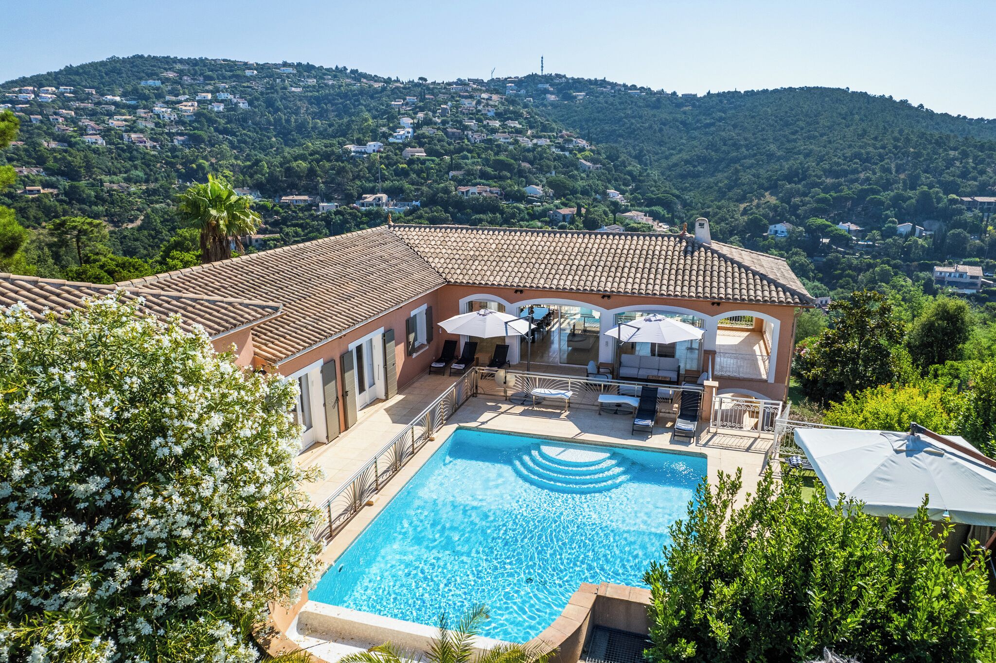Une villa dotée d’une piscine, d’un espace extérieur aménagé et offrant une vue sur une colline parsemée de maisons.