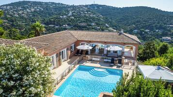 Une villa dotée d’une piscine, d’un espace extérieur aménagé et offrant une vue sur une colline parsemée de maisons.