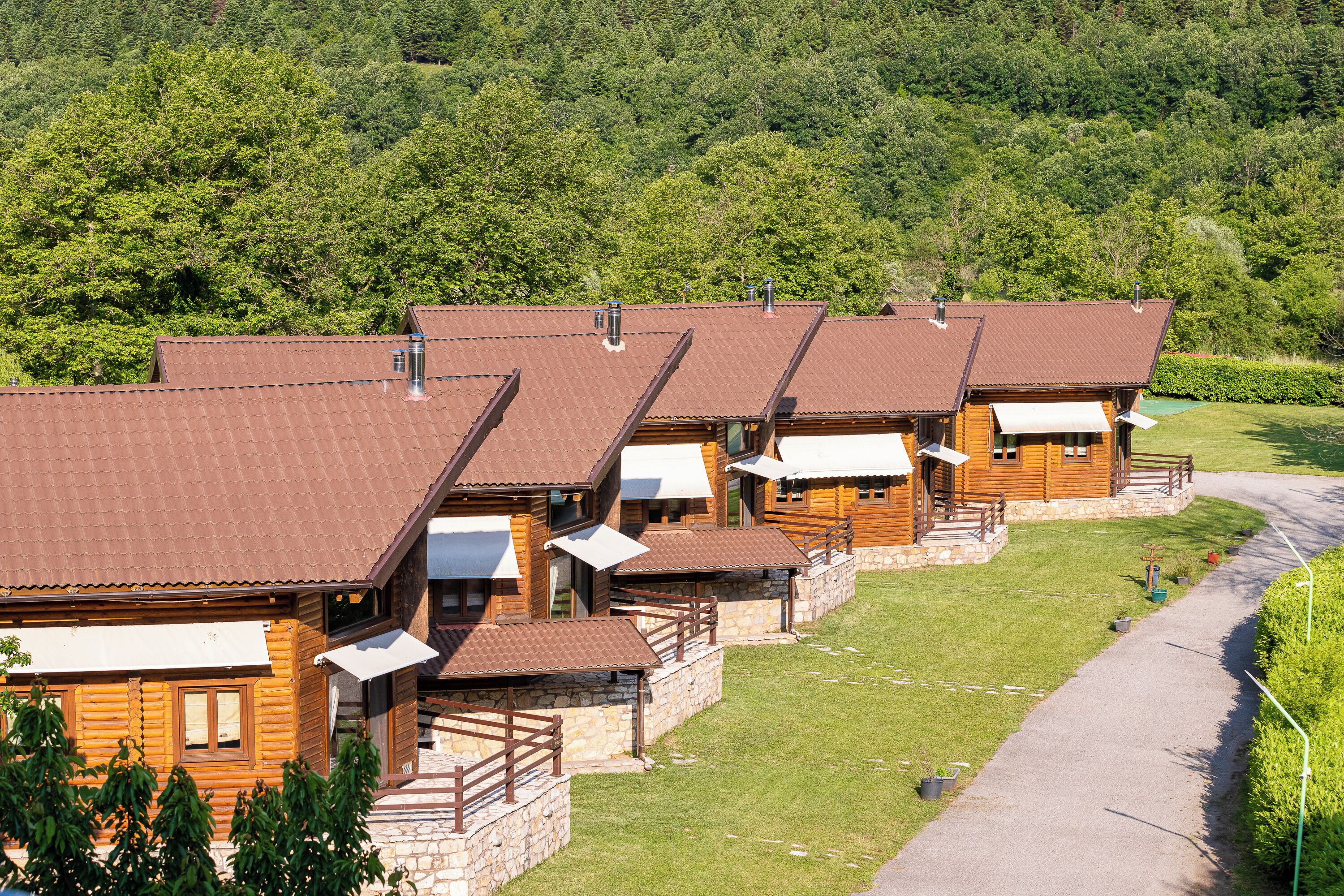 A row of wooden cabins with sloped roofs, arranged in a linear fashion along a paved path, surrounded by lush greenery and a forested backdrop.