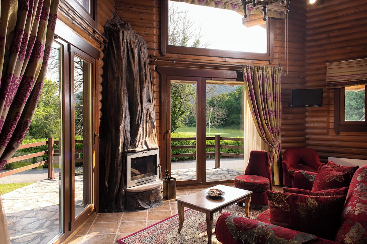 A log cabin interior with a stone fireplace, large windows, and a red upholstered sofa.