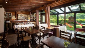 A rustic wooden interior with a dining area, kitchen, and large windows overlooking a playground. - Natura Chalets (Karpenisi)