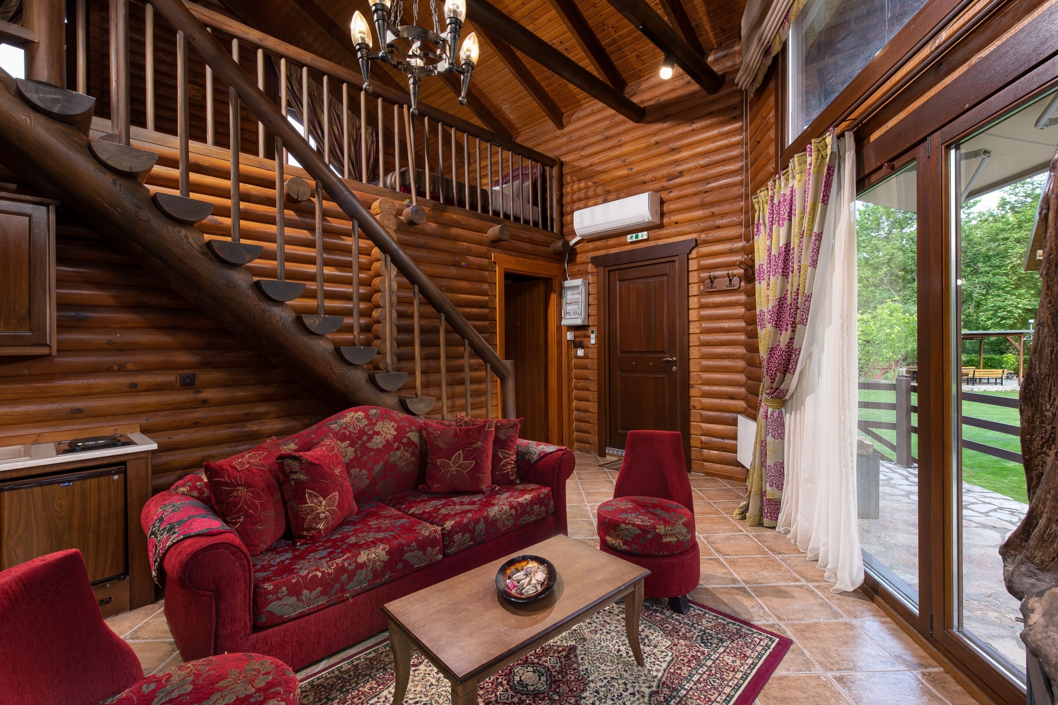 Interior de una cabaña de troncos con una escalera de madera, un sofá rojo, una mesita y vistas del exterior a través de grandes ventanales.