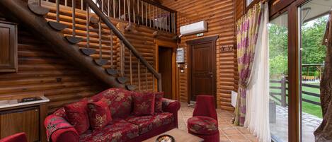 A log cabin interior with a wooden staircase, a red sofa, a small table, and a view of the outdoors through large windows.