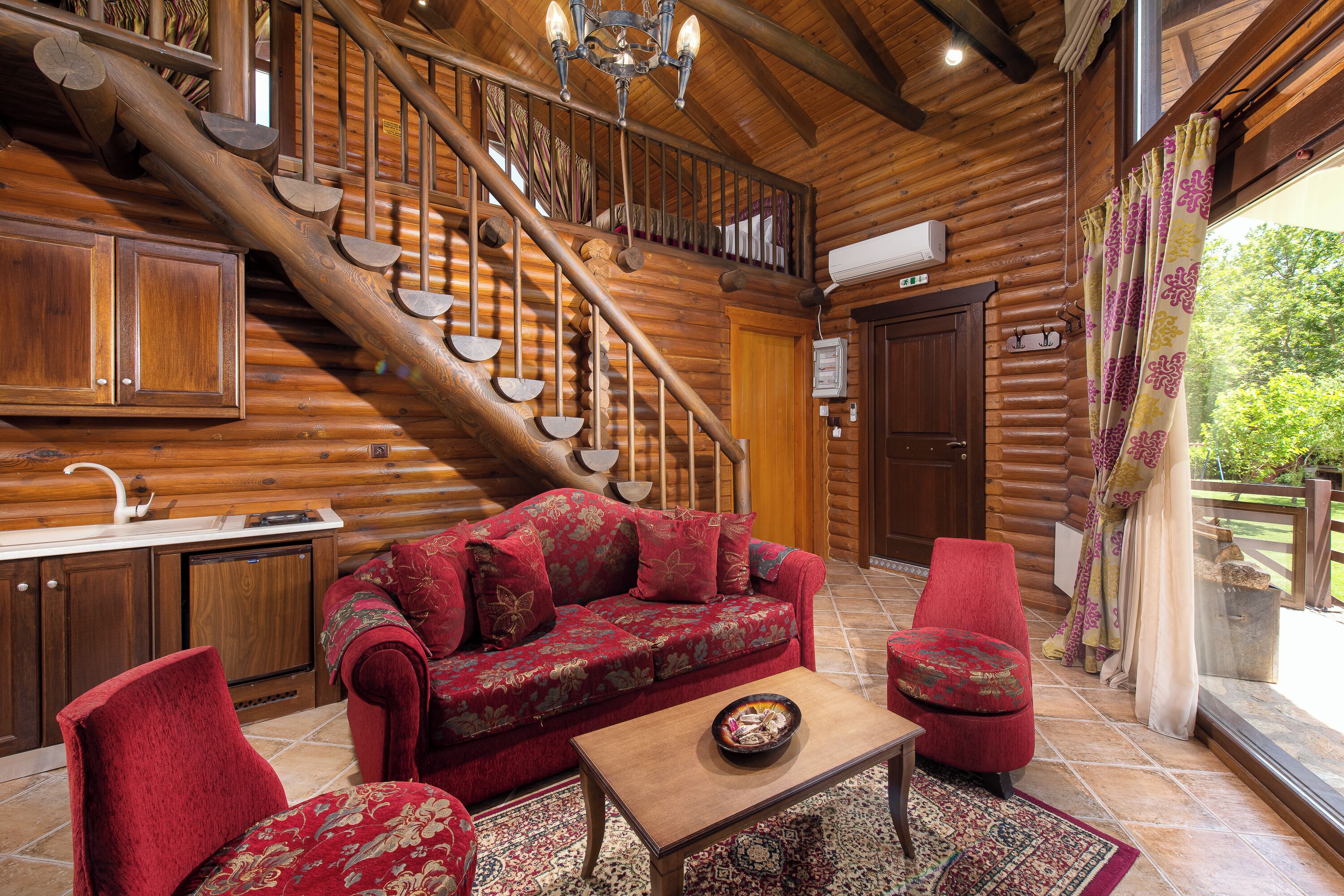 A log cabin interior with a wooden staircase, a red sofa, a wooden coffee table, and a view of the outdoors through a window.