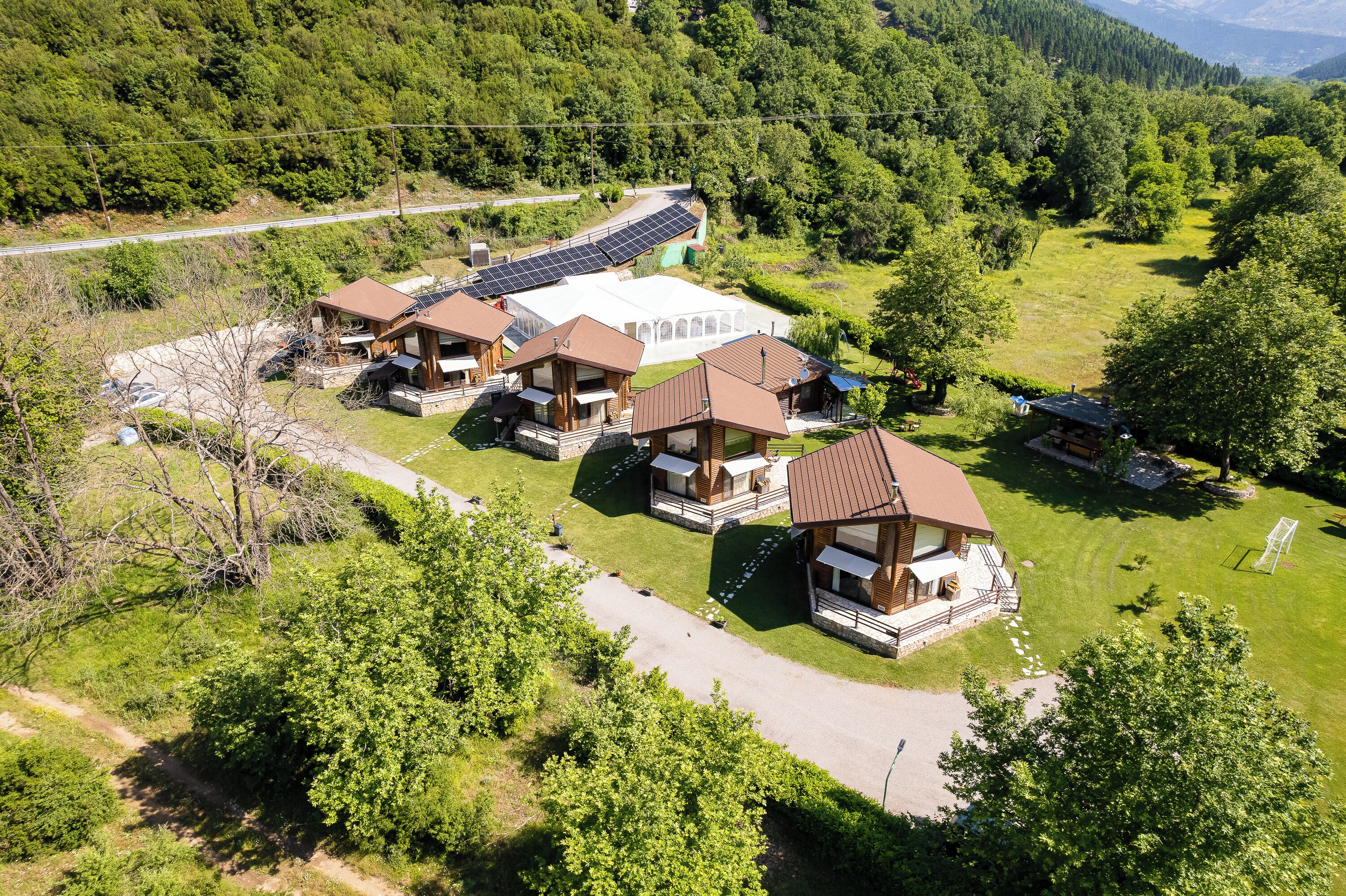 A cluster of wooden cabins with solar panels, surrounded by lush greenery and a mountain backdrop.