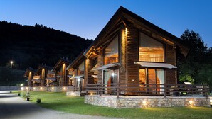 A row of log cabin-style lodges with stone foundations and wooden railings, set against a mountainous backdrop. - Natura Chalets (Karpenisi)