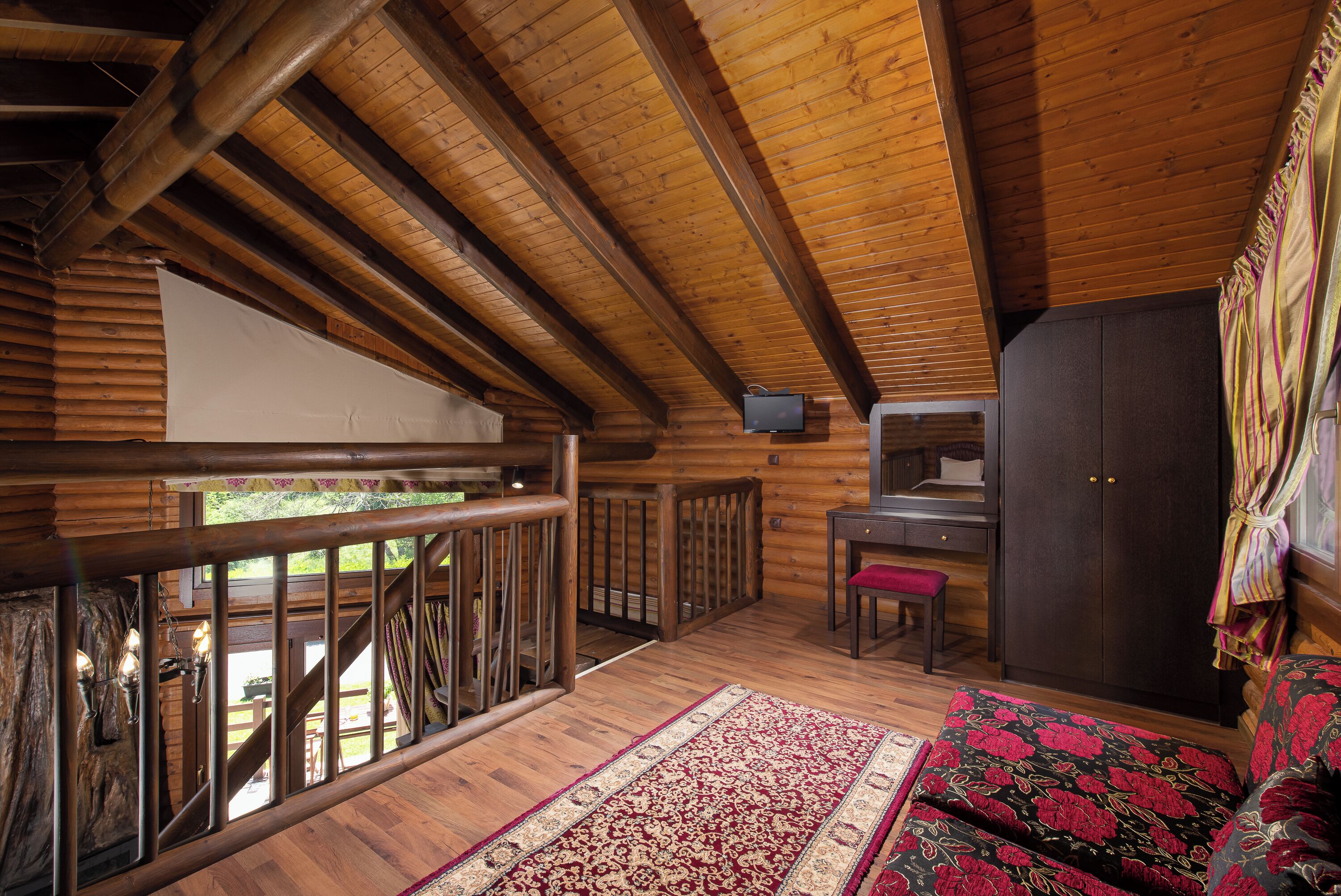 A wooden cabin interior with a balcony, a desk, a wardrobe, and a patterned rug.