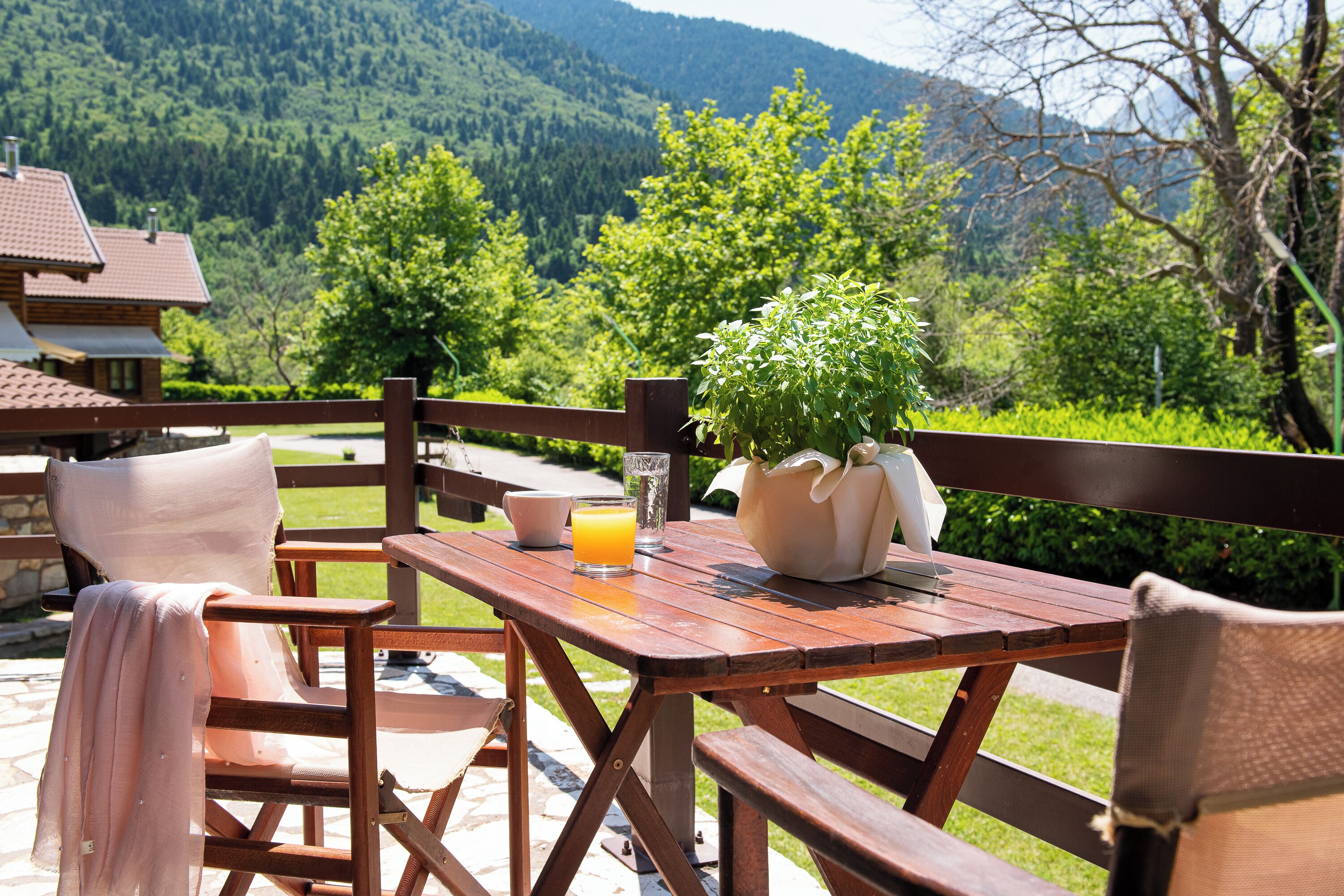 A wooden table and chairs set outdoors with a mountain view, a glass of orange juice, and a potted plant.