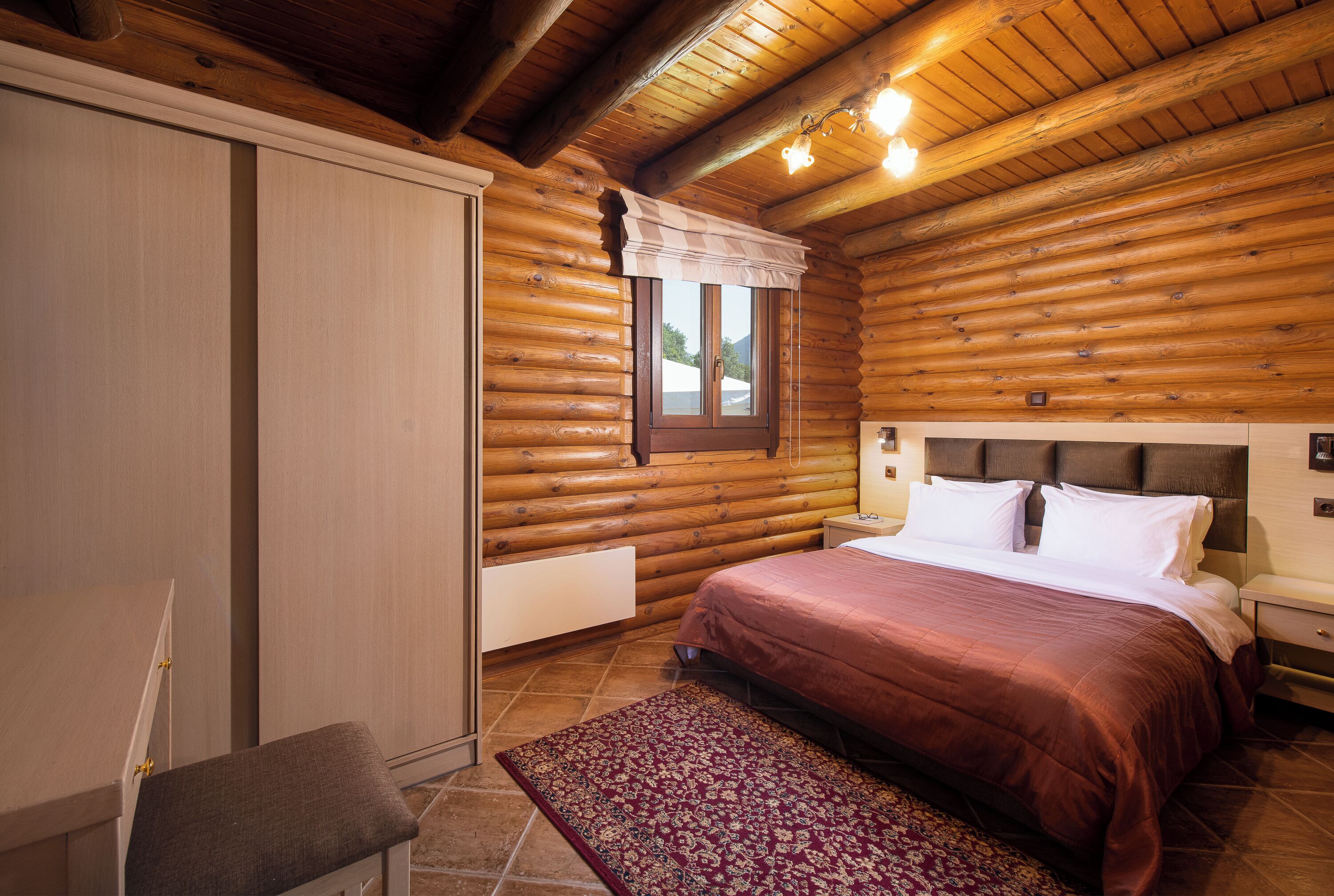 Ein Schlafzimmer in einer Blockhütte mit einem Bett, Nachttischen und einem Fenster mit weißem Vorhang.
