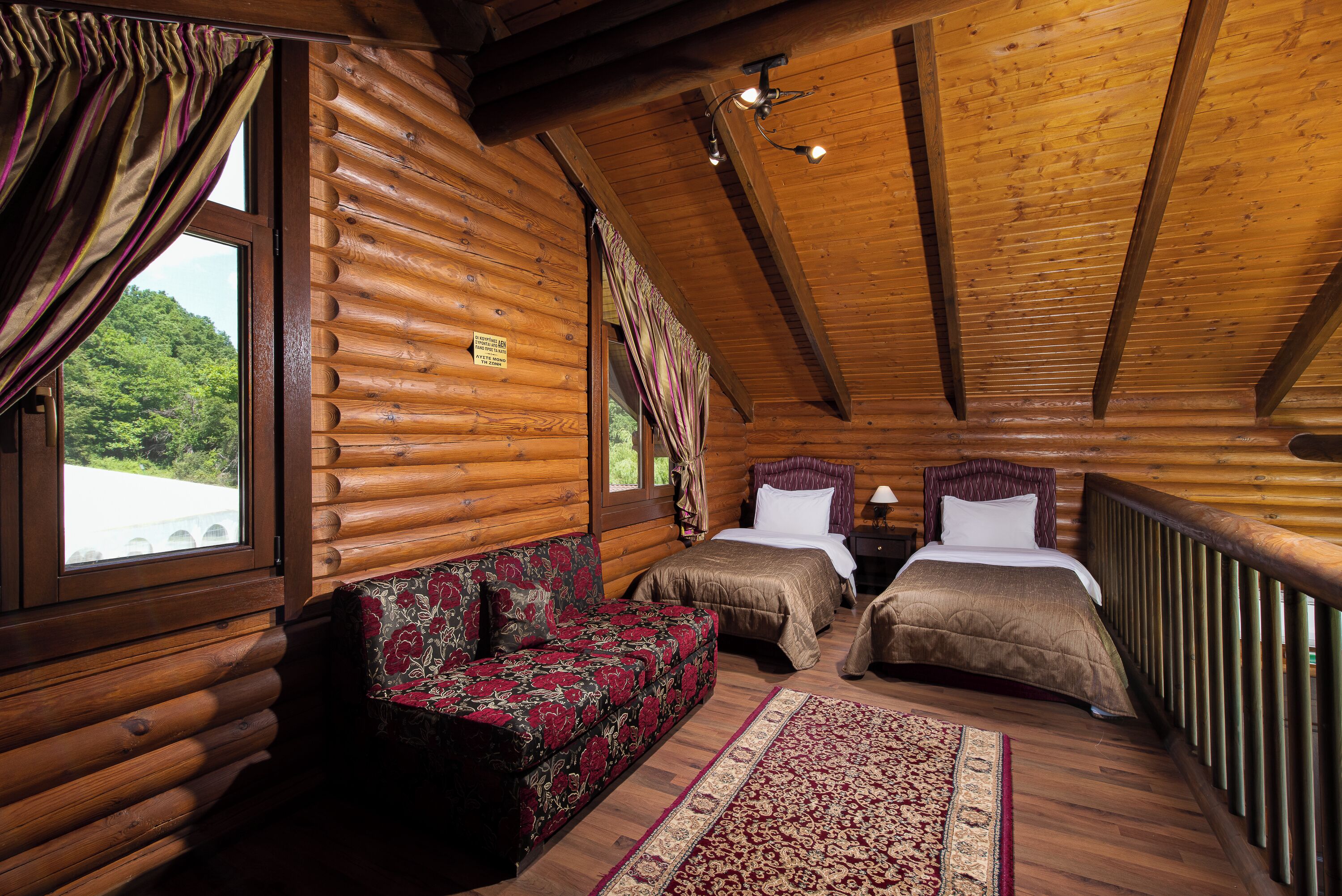 A log cabin bedroom with two beds, a sofa, and a view of a forest.