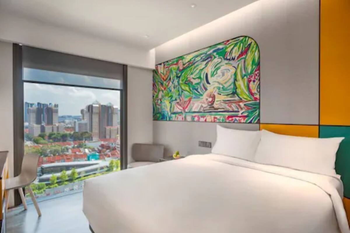 A modern hotel room with a large bed, a city view, and a colorful abstract painting on the wall.