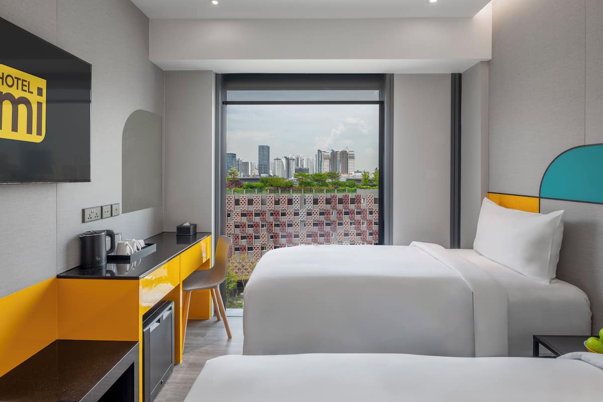A hotel room with a bed, a desk with a kettle, and a view of the city skyline.