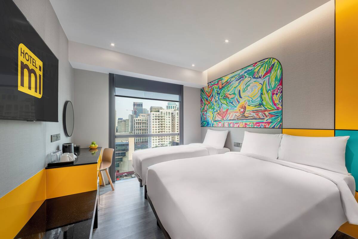 A hotel room with two beds, a large window with a city view, a colorful abstract painting on the wall, and a minibar.