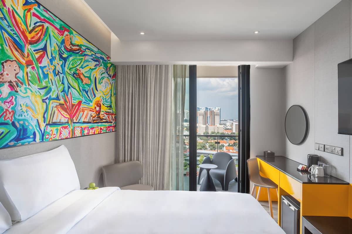 A hotel room with a bed, a desk, a chair, a small table, and a colorful abstract painting on the wall.