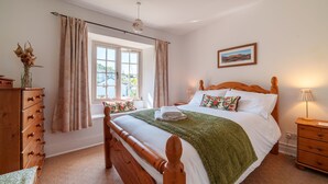 A bedroom with a wooden bed, a window with curtains, a bedside table with a lamp, and a dresser. - Red Robin Cottage -  Garden, Parking and Walk to the Lake (Ambleside)