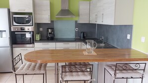 Een compacte keuken met een ingebouwde oven, magnetron en koffiezetapparaat. Er staat een houten tafel met vier stoelen en een glazen kan op.