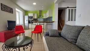 Een moderne woonkamer met een grijze bank, rode stoelen, een glazen salontafel en een rode entertainmentkast. Achtergrond toont een keuken met witte kasten en een groene accentmuur.